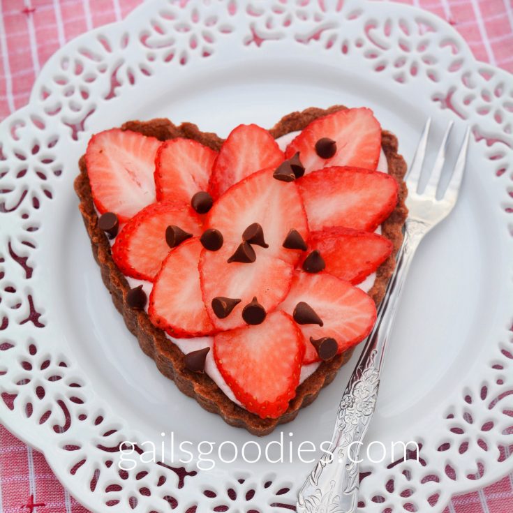 A single strawberry marshmallow tart on a white dessert plate with lacey edges. The heart-shaped tart is topped with fresh strawberries and sprinkled with mini chocolate chips. A little of the pink strawberry marshmallow mousse peeks out between the fresh strawberries and the chocolate crust. There is a fork on the plate to the right of the tart.