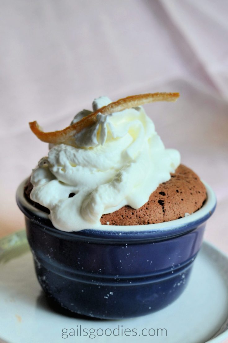 A dark chocolate grand marnier souffle in a blue ramekin. The chocolate souffle mounds ovr the top of the ramekin. The souffle is topped with whipped cream and a small piece of candied orange peel.