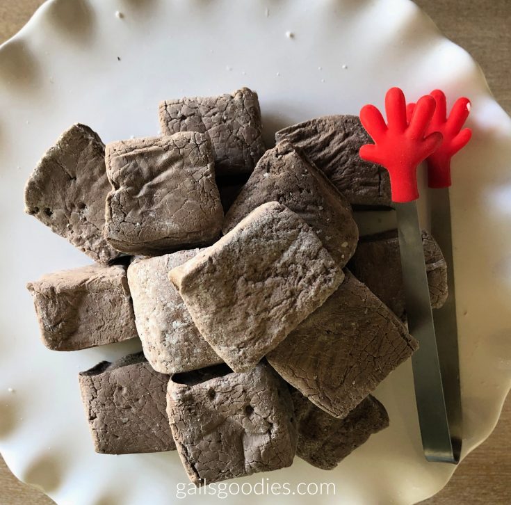 A stack of chocolate marshmallows viewed from the top. The marshmallows are cut into small rectangles and stacked in a pyramid on a white plate. A pair of tongs with red hands is on the right side of the plate.