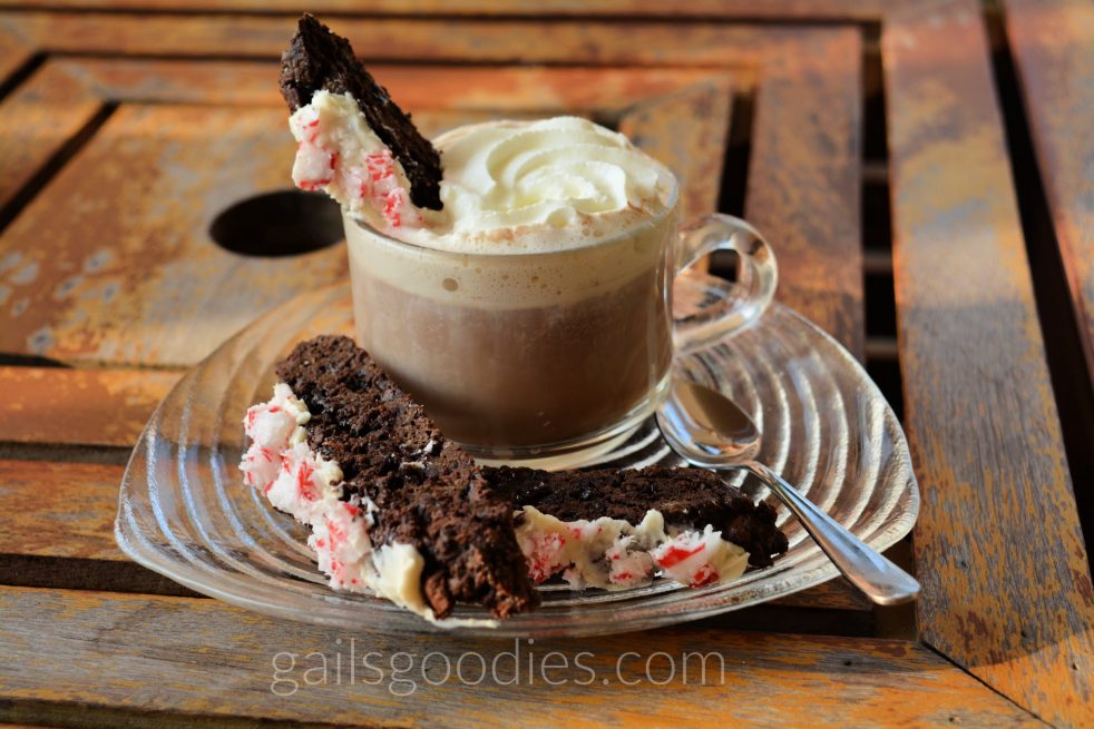 Hot chocolate with whipped cream in a glass mug that sits on a glass plate. Two piece of chocolate peppermint biscotti are to the left of the mug and a spoon is to the right of the mug. A piece of chocolate peppermint biscotti rises out of the hot chocolate on the left.