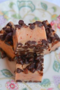 This is the top view of four cubes of Chocolte Covered strawberry fudge on a floral plate. There are three pieces on the bottom and one on top. The top piece is turned to the side. There are mini chocolate chips on each side and pink strawberry fudge in the middle.