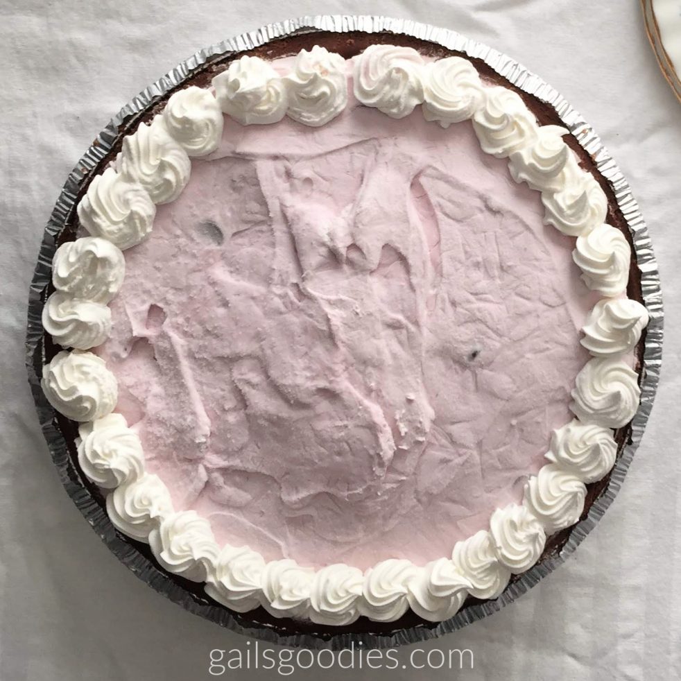 A whole black forest yogurt pie is viewed from above. The medium pink filling is circled by swirls of whipped cream and the dark chocolate crust peeks out along the outer edge of the pie.