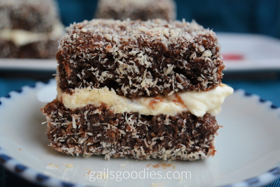 A close up of a single easy lamington viewed from the side. The bottom and top layers are dark chocolate speckled with white cocolnut flakes. The middle layer is creamy whipped cream. Some of the chocolate from the top layer has spread into the whipped cream in the middle right of the cake.