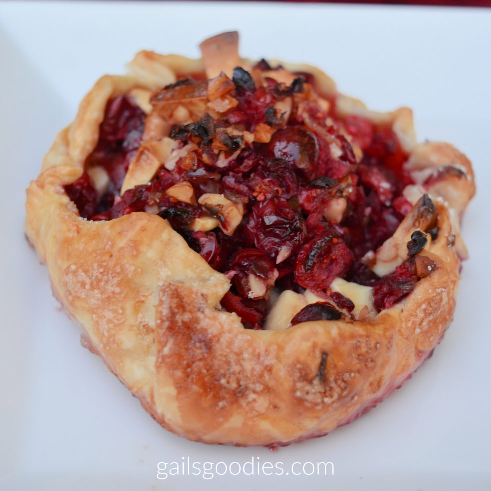 A cranberry walnut galette sits on a white plate. The golden brown pastry edges are folded up around the bright red cranberry filling. Light brown pieces of walnut are scattered throughout the filling.