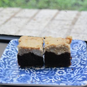 Two brownies sit on a blue paisley plate. The dark chocolate brownie base contrasts sharply with the pale brown mocha cheesecake topping.  
