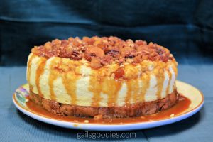 A creamy pale yellow cheesecake sits on top of a one inch thick light brown blondie. The cake is topped with candied nuts and caramel sauce. The caramel sauce drips down the sides of the cake so the sides of the cake are striped light brown caramel and pale yellow cheesecake.