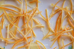 Candied citrus peel spread out on a white silicone mat. The candied peel is cut into thin strips that are dark orange on one side and pale yellow on the other. 