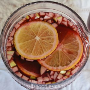 The top view of a pitcher of Macanese sangria. There are two orange rounds and finely diced green apple floating in the dark raspberry colored liquid. 