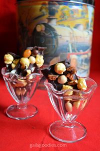 Two glass ice-creamdishes filled with pieces of mocha caramel popcorn bark sit on a red tablecloth. There is a decorative tin in the background. The dark chocolate bark is irregularly shaped and dotted with pieces of golden caramel corn, medium brown and pale brown M&Ms and pecan pieces.