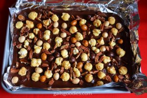 Mocha caramel popcorn bark in a foil lined pan. the dark chocolate is dotted with pieces of golden caramel popcorn, light brown and pale brown M&Ms and pecans.