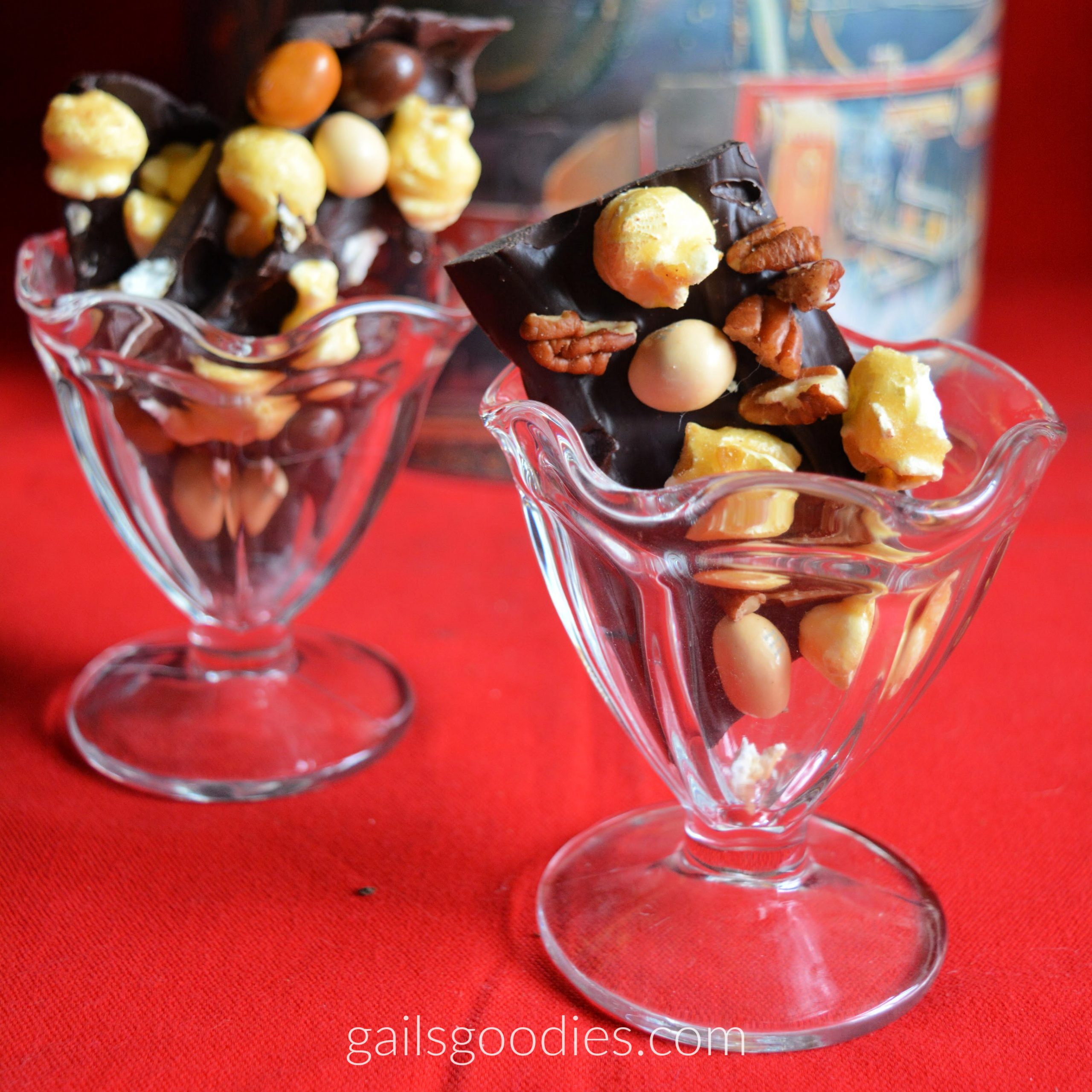 Two glass sundae bowls hold pieces of mocha caramel popcorn bark. The dark chocolate bark pieces are irregular shapes and are dotted with pieces of golden caramel corn, medium and pale brown m&Ms and pecans. The glasses sit on a red tablecloth and there is a decorative tin in the background.