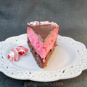 A slice of cheesecake sits on a white plate with lacey edges. The point of the slice is at the front of the photo. The brownie bottom is topped with red peppermint cheesecake. The slice is topped with a thin layer of dark chocolate ganache and there are crushed peppermint candies sprinkled on the outer edge of the slice. Two pillow peppermint candies sit on the plate to the left of the slice.