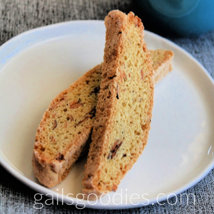 Two slices of almond biscotti on a round plate. The slice on the left is flat on the plate and the slice on the right is angled on top of the left one about halfway back. The golden biscotti is dotted with almond slivers.