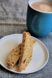 Two slices of almond biscotti are on a white plate. The two slices criss cross at the back with the slice on the right is on top. There is a blue mug of coffee in the upper right corner of the photo