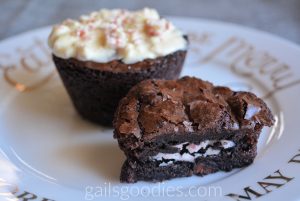 Two peppermint oreo stuffed brownies on a plate. The one in back is muffin shaped and topped with white chocolate and curshed peppermint. The one in front has been cut in half and the cut side faces front revealing peppermint oreo in the middle on the brownie.