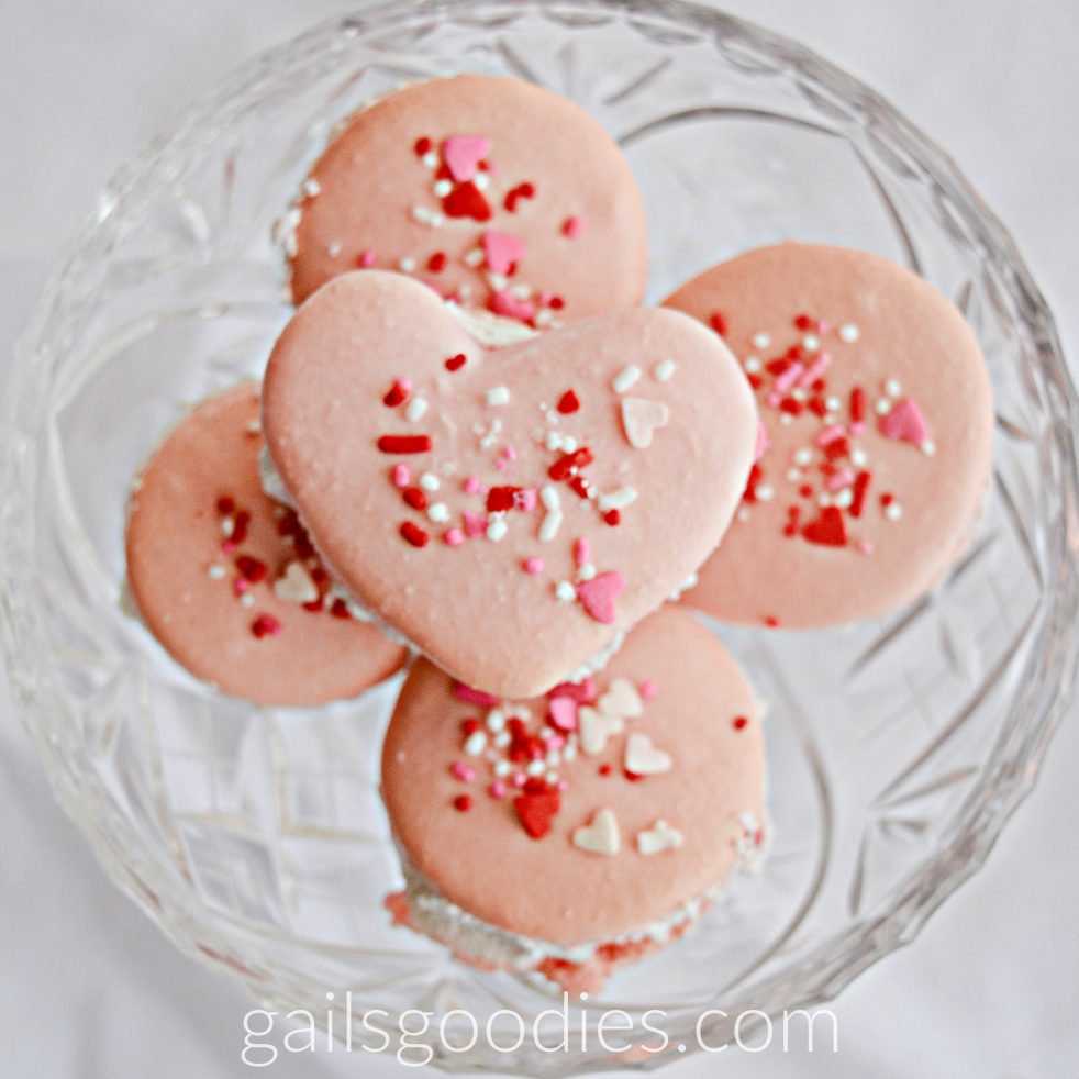Five pink macarons in a cut glass dish. The four on the bottom are round and the one on top is heart shaped. The macarons are topped with red and white sprinkles.
