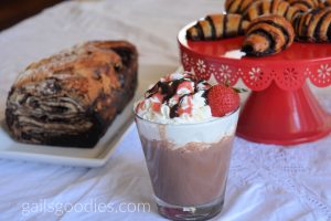 A glass of strawberry hot chocolate is in the front center of the photo. The hot chocolate is topped with whipped cream drizzled with chocolate syrup and strawberry syrup. There is a frshe strawberry on the right side of the glass.  Behind the glass to the right is a red cake stand with golden chocolate rugelach. Behind the glass on the left is a load of chocolate babka.