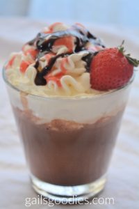 A clear glass filled with strawberry hot chocolate. The hot chocolate is topped with whipped cream drizzled with chocolate sauce and strawberry sauce. There is a single fresh strawberry garnishing the glass on the right.
