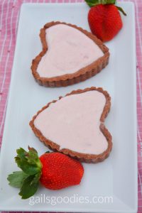 Two heart shaped tarts are on a rectangular white tray. They face in opposite directions. The chocolate pastry is filled with pink strawberry marshmallow mousse. There are fresh strawberries at each end of the tray.