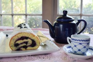 This is a photo of a table set for tea. On the left, there is a rectangular white platter with a vanilla blueberry cake roll. The cut end is facing front revealing a spiral of vanilla sponge with blueberry filling. Just left of center there is a dark blue tea pot. There is a blue and white tea cup in the bottom right corner of the photo.