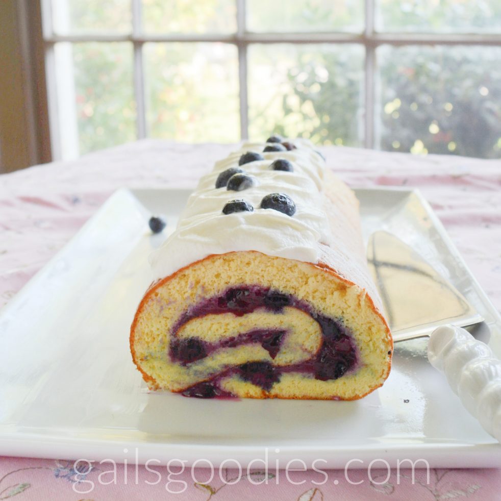 A vanilla bluberry cake roll sits on a rectangular white platter. The cut end faces front revealing a swirl of vanilla cake with blueberry filling. The cake roll is topped with whippd cream and fresh blueberries.