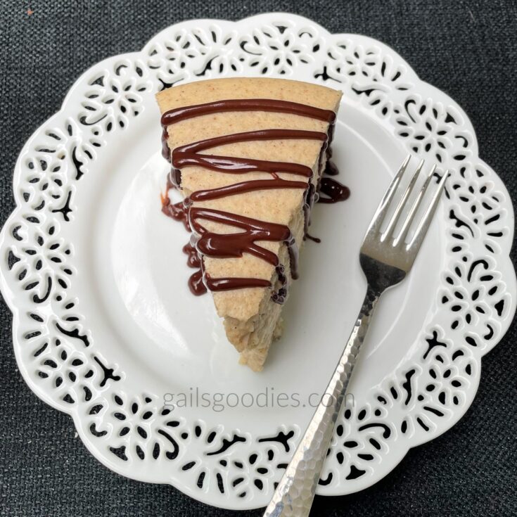 This is a top view of chai spice cheesecake. The tan colored cheesecake is drizzled with dark chocolate ganache. There is a fork on the plate to the right of the slice.