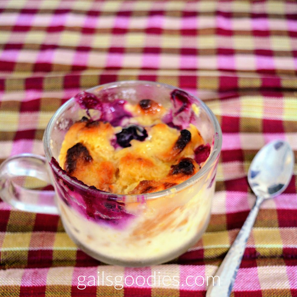 Lemon Blueberry bread pudding served in a glass teacup. The top is golden brown and blueberries peak through between the bread cubes. The sides of the cup have purple blueberry juice on top and creamy custard on the bottom. A spoon sits on the table to the right of the cup