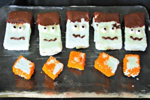Five light green, rectangular lime marshmallows are arranged in a row. Each marshmallow is decorated as Frankenstein. The faces are made from two candy eyes and a squiggly chocolate mouth. The tops of each marshmallow are dipped in chocolate to make hair. There are five cube-shaped marshmallows in a row below the frankenstein marshmallows. The cube-shaped marshmallows have orange nonpareils on some sides but not others.