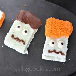 The pale green marshmallow on the right is rectangular in shape. It is decorated with orange nonpareils as hair. Two candy eyes and a chocolate squiggle mouth make the face. The light green marshmallow to the left is also rectangular. It has a face made from two candy eyes and a squiggly chocolate mouth and the top is dipped in chocolate to represent hair. 