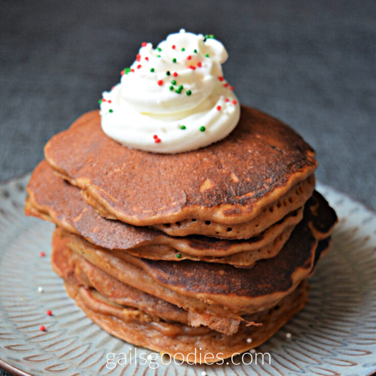 A closeup of the stack of pancakes. There are about 8 pancakes in the stack. The pancakes are medium brown on top and tinged orange on the edges. There is a swirl of whipped cream on top and the whipped cream is dotted with green, red and white sprinkles.