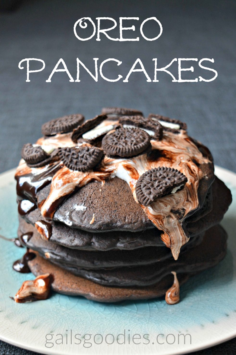 A stack of 5 black chocolate oreo pancakes sit on a teal plate. The pancakes are topped with marshmallow cream, fudge sauce and broken mini-oreos. The words "oreo pancakes" are at the top of the photo.