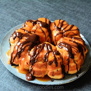 This is a photo of the whole loaf of monky bread. The ring-shaped loaf has small segments than can be pulled apart. The golden loaf is topped with drizzles of chocolate sauce and caramel sauce.