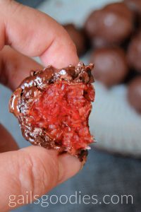 This is a close up shot of a cherry ripe truffle with a bite taken out of it. The bright red coconut cherry filling is surrounded by milk chocolate. There is an out of focus plate of more cherry ripe truffles in the background.