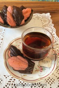 There is a plate with two madeleines and a glass mug of tea in the front of the photo. There is a bowl of chocolate cherry madeleines in the background of the photo.