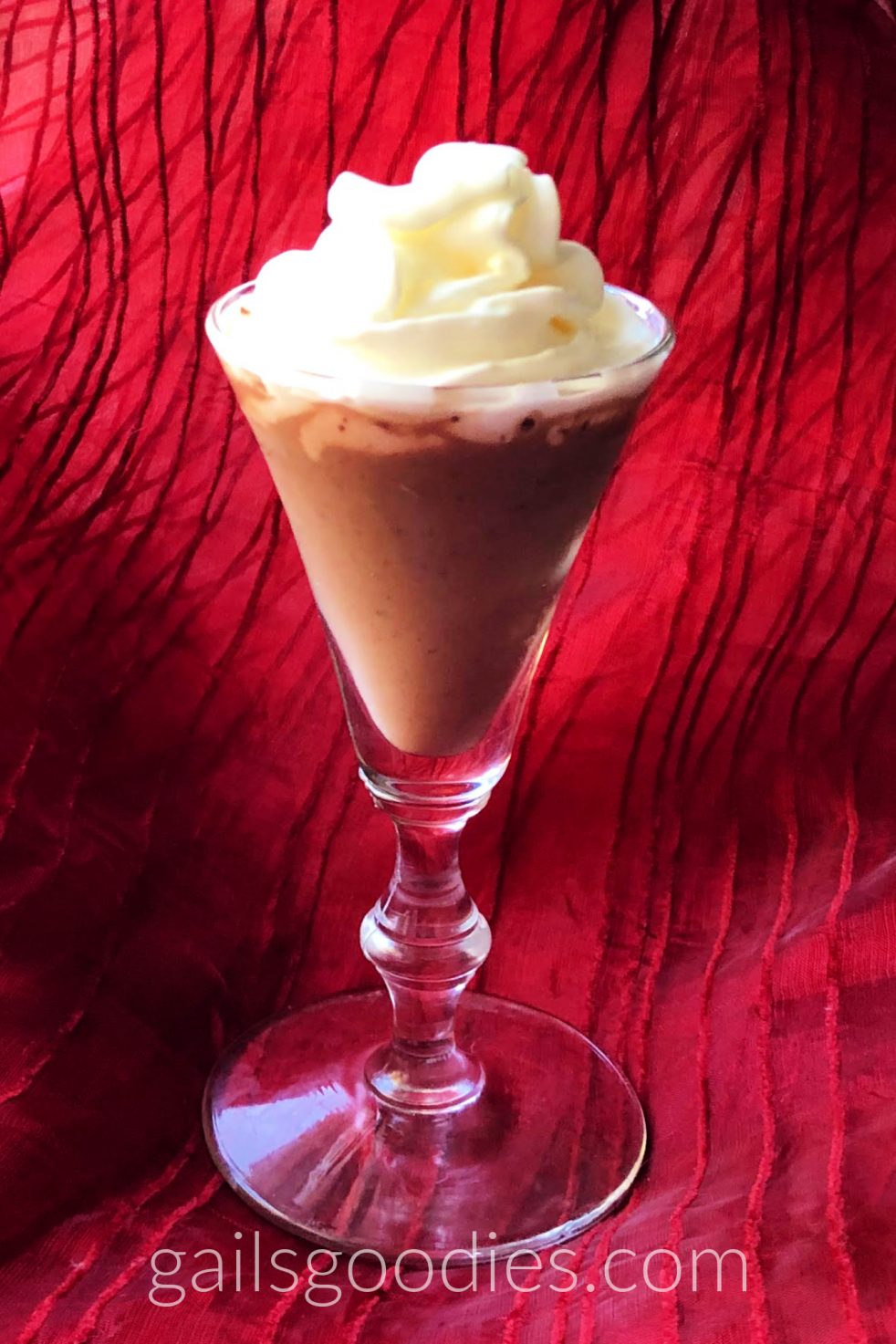 A plain stemmed liqueur glass is filled with a milk chocolate colored liqueur. Whipeed cream is swirled on the top and the photo has a red organza background and is lit from the left side.