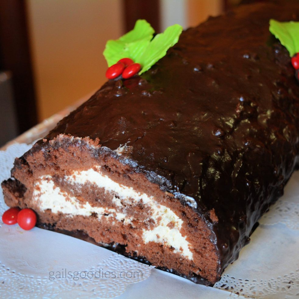 Chocolate cake spiral with whipped cream covered with chocolate ganache. The chocolate roll cake is garnished with green chocolate leaves and round red candies in groups of three to replicate the look of holly and berries.