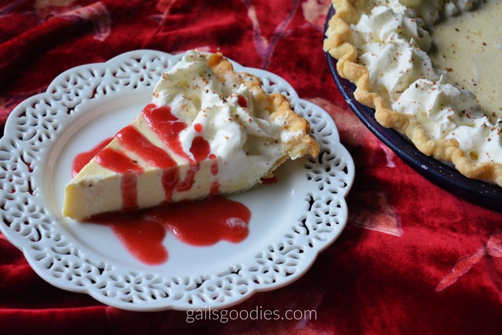 A slice of creamy eggnog pie sits on a white dessert plate with a lacey edge. Swirls of whipped cream are dotted along the edge next to the crust. The slice is drizzled with raspberry sauce and ground nutmeg is sprinkled on the whipped cream. The edge of the whole pie is in the upper right corner.