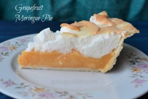 Slice of grapefruit meringue pie sits on a white plate with pink and orange flowers on the border. The peachy orange filling sits ona flakey crust and is topped with an equal amount of fluffy meringue.