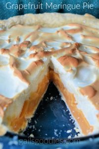 Grapefruit mering pie in a blue pie plate. The background is teal and blue. There is a slice removed from the front so the peach colored filling is visible under the fluffy meringue topping.