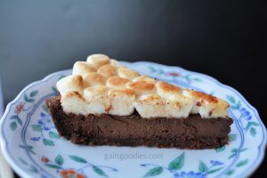 A small slice of a dark chocolate tart with chocolate crust and toasted marshmallow topping sits on a white plate with a blue and orange floral border.