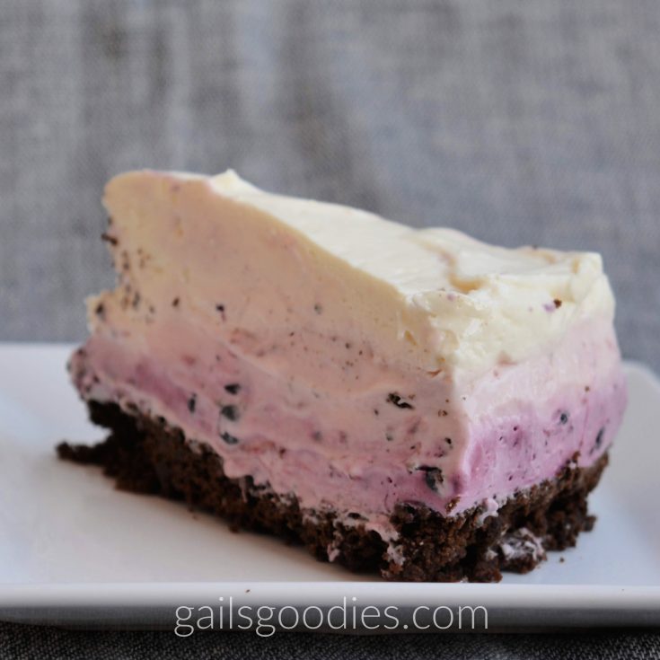 A slice of ombre berry cheesecake sits on a square white plate. The bottom layer is a dark chocolate crust, followed by a purple blueberry layer, then a pink strawberry layer and finally a creamy white vanilla cheesecake layer.