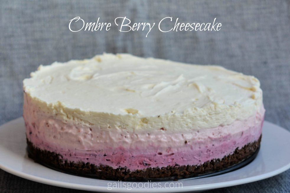 The photo shows a whole cheesecake on a plate. The dark chocolate crust is topped with layers of blueberry, strawberry and vanilla cheesecake. The bottom blueberry layer is a dark purple and the strawberry layer is a light pink. The vanilla layer on top is a creamy white. The background is grey and the words "Ombre Berry Cheesecake" are at the top of the photo.