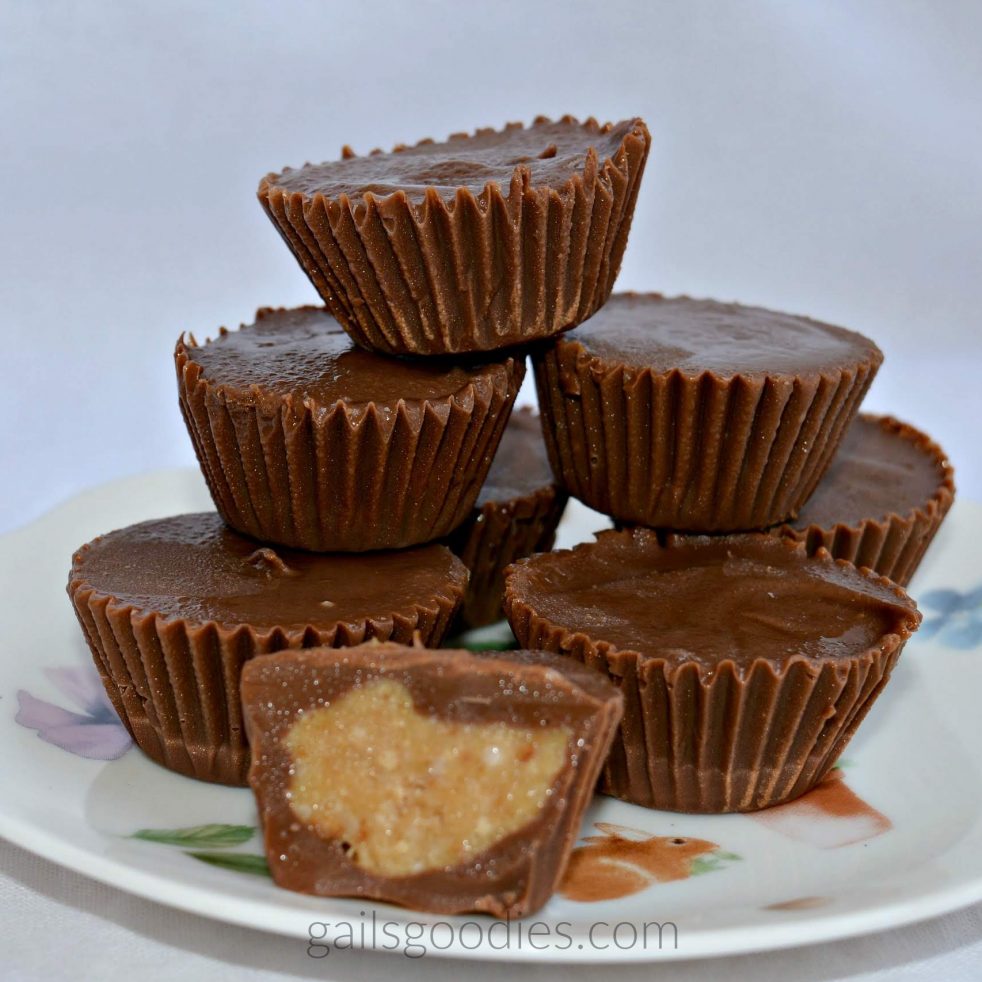 Eight chocolate peanut butter cups are stacked in a pyramid on a plate. A peanut butter cup cut in half sits in front of the stack. The filling is creamy with darker brown flecks.