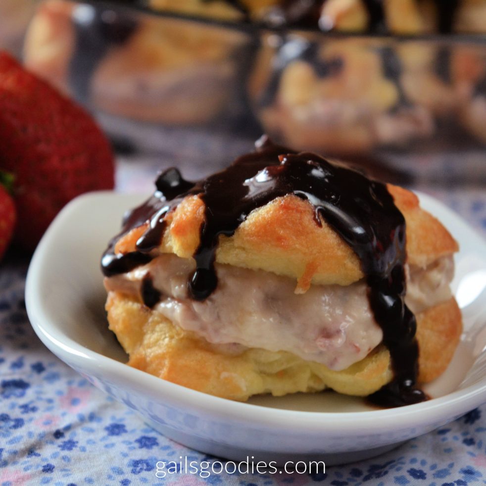 A single strawberry cream puff sits on a small white plate. The golden choux pastry is cut open horizontally revealing the creamy strawberry filling. Dark Chocolate ganache coveres the top and is dripping down the sides. To the left of the plate are some fresh strawberries.