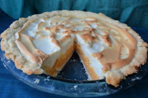 Grapefruit mering pie in a blue pie plate. The background is teal and blue. There is a slice removed from the front so the peach colored filling is visible under the fluffy meringue topping.