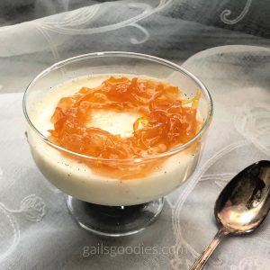 A single stemmed glass filled with pale yellow panna cotta sits on a lace cloth. The panna cotta has tiny black flecks from vanilla bean seeds and is topped with orange-colored marmalade. There is a silver spoon to the bottom right of the glass.