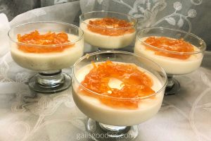 Four stemmed glasses are filled halfway with a pale yellow panna cotta. Little black flecks of vanilla bean seeds dot the pale yellow dessert and there is a circle of orange colored marmalade on top of each dessert.