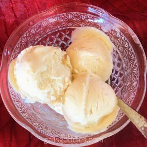This is a top view or a bowl of lemon curd ice-cream. Three scoops of cream yellow lemon curd ice-cream are in a cut glass bowl. In the lower right portion of the bowl there is a spoon emerging from the ice-cream. The bowl is on a red organza cloth.