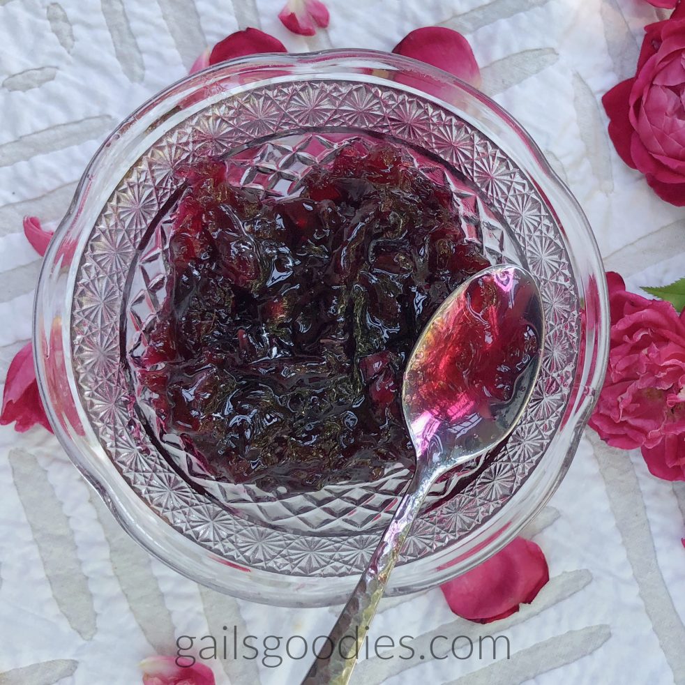 This is a view from above of a cut glass bowl with rose jelly. The deep magenta rose jelly is textured with rose petals. There is a silver spoon on the right side of the container. Some of the jelly fills the bowl of the spoon. There are magenta roses on the white table cloth to the right of the bowl and magenta rose petals are scattered on the tablecloth around the glass bowl.