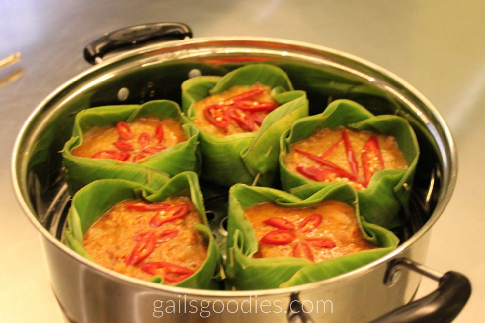 Five banana leaf bowls filled with orange fish amok.They are in a pan and each is decorated with slices of thai chili.